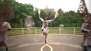 Three dancers in flowing dresses perform a traditional dance on an outdoor stage with a lush green backdrop and a historic building video