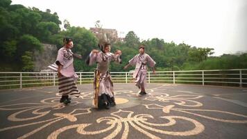 Traditional dancers in vibrant costumes performing outdoors with a scenic backdrop of lush greenery and a distant building video