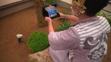 Person in traditional attire taking a photo of a plant with a tablet in a greenhouse setting. video