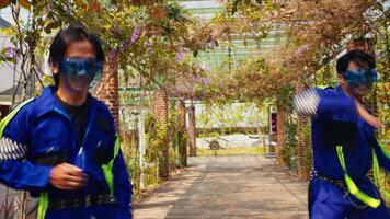 Two people in blue overalls and safety goggles playfully dancing in a garden with a vine-covered archway. video