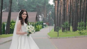 The bride walks alone in the park and turns to the camera and smiles. Wedding day and wedding walk. The bride turns to the camera, smiles, holds a wedding bouquet of white roses in her hands. video