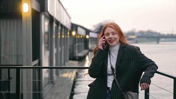 Woman talking on phone by waterfront, A woman with red hair and a striped blazer is talking on her phone while standing by a waterfront. video