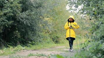 A girl in a yellow raincoat stands in the forest on the road and uses a mobile phone video