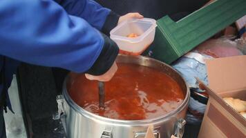 portion soupe dans une dans une Plastique récipient, une la personne dans une bleu veste louches soupe dans une récipient de une grand pot à une communauté cuisine, mise en évidence le acte de fournir chaud repas à ceux dans besoin. video