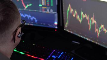 Exchange stock market. Back view of young professional trader in eyeglasses pointing on the data on computer screen with pen and holding Bitcoin in one hand while working his modern office video