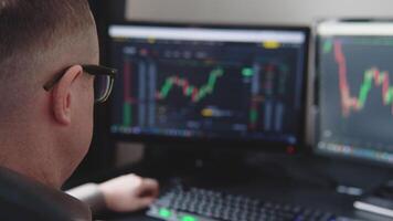 Trader wearing eyeglasses sitting at desk at office monitoring cryptocurrency chart looking at monitors analyzing candle bar price flow touching chin concerned trading concept close-up video