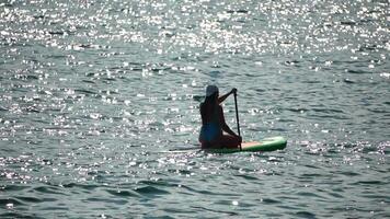 mar mujer sorber. silueta de contento positivo joven mujer en azul bikini, surf en verde cenar tablero mediante calma agua superficie. idílico puesta de sol. activo estilo de vida a mar o río. lento movimiento video