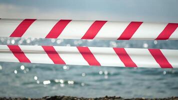 rood wit waarschuwing plakband barrière lint swinging in de wind aan de overkant exotisch zee strand achtergrond zonder mensen. Nee binnenkomst rood wit voorzichtigheid plakband. Nee vakantie concept, vertraagd reis, Nee zomer plannen video