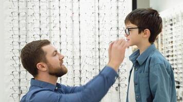 Little boy in optics store choosing glasses with his father. Ophthalmology concept video