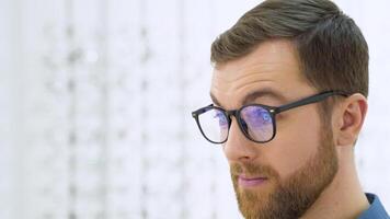 Portrait of handsome bearded guy picking new glasses at optical shop, looking at mirror video