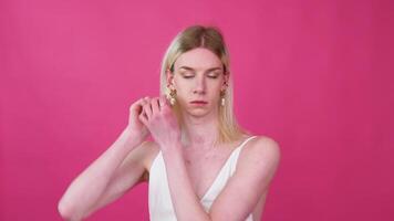 Portrait of young transgender man in a dress puts on earrings on a pink background video