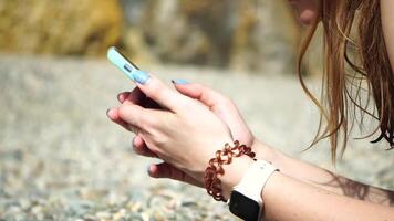 Woman with smartphone. Close-up of woman's hands holding vertical mobile phone and swiping up finger application page against background of sea and beach video