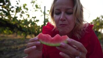 Woman eat watermelon. Close-up Portrait of a cheerful blond woman in red dress eating slice of watermelon and smiling at camera. Happy lady enjoy taste of juicy fruit outside in park. Slow motion video