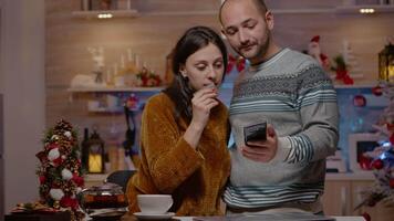 feestelijk paar buying presenteert met credit kaart Aan smartphone, aan het doen online boodschappen doen voor Kerstmis vooravond viering. mensen gebruik makend van apparaat betalen voor cadeaus in seizoensgebonden versierd keuken video