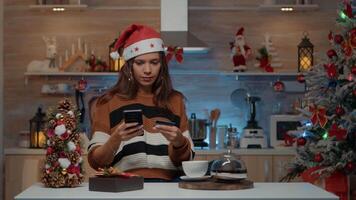 festivo adulto utilizando teléfono inteligente para en línea compras con crédito tarjeta en estacional decorado cocina. mujer con sombrero consiguiendo Navidad regalos para celebracion cena fiesta con amigos video