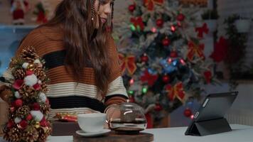 Woman offering gifts on videocall using modern tablet at home in christmas designed kitchen. Festive young adult with sweater sharing present for seasonal celebration festivity greeting video