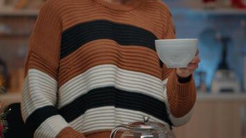 Close up of young adult holding cup of tea in kitchen with utensils designed for festive christmas eve celebration party at home. Woman preparing winter beverage from kettle on counter video