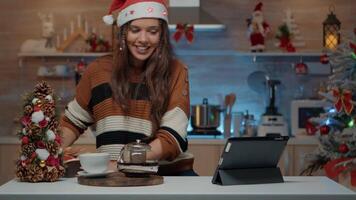 sorridente mulher usando ligar tecnologia para apresenta dentro festivo decorado cozinha com árvore e enfeites. pessoa com santa chapéu dando virtual presentes para família em Natal véspera dia video