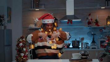 leende kvinna med santa hatt tilltrasslad i jul lampor sträng medan håller på med dekor förberedelser i kök. ung person har roligt inomhus med festlig dekorationer och ornament video