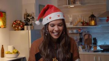 Festive woman with santa hat receiving gift on call from family in holiday decorated kitchen at home. Young adult being happy while opening present for christmas eve festivity video