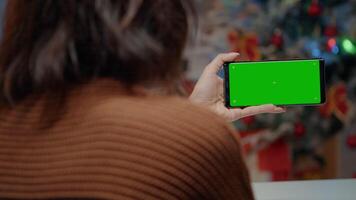 Young woman using green screen smartphone in festive kitchen decorated for christmas. Adult holding chroma key background device for digital mock up template and modern technology video