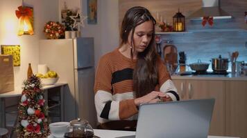 caucásico mujer sentado a cocina mesa con ordenador portátil esperando para invitados a llegar para Navidad cena fiesta a festivo decorado hogar. triste adulto comprobación reloj para amigos y parientes video