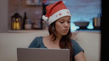 Festive person working with laptop and watching television on christmas holiday. Woman with santa hat looking at device and TV screen while eating chips on winter celebration video