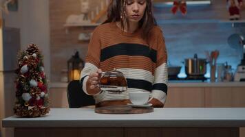 Caucasian woman sitting in christmas decorated kitchen pouring tea from kettle. Young person getting warm with winter beverage while preparing for seasonal holiday celebration party video