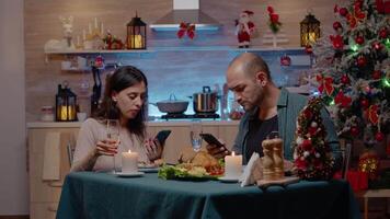 moderno Pareja utilizando teléfonos inteligentes disfrutando festivo cena en Navidad víspera. hombre y mujer mirando a dispositivos mientras sentado a mesa con comida y champán para fiesta celebracion. video