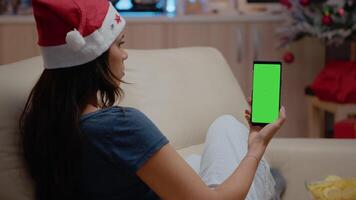 Woman vertically holding smartphone with green screen on christmas eve. Person looking at mockup template and isolated background on cell phone device with chroma key. Adult with gadget video