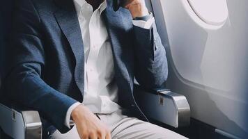 geslaagd Aziatisch zakenman in pak en bril zit in privaat Jet en toepassingen smartphone, Koreaans ondernemer vliegt in een vliegtuig en types Aan de telefoon online video