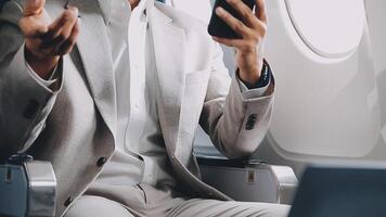 successful asian businessman in suit and glasses sits in private jet and uses smartphone, korean entrepreneur flies in an airplane and types on the phone online video