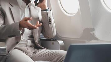 framgångsrik asiatisk affärsman i kostym och glasögon sitter i privat jet och användningar smartphone, koreanska entreprenör flugor i ett flygplan och typer på de telefon uppkopplad video