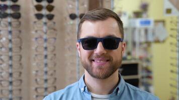 Portrait of handsome bearded guy picking new sunglasses at optical shop video