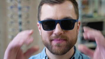 Portrait of handsome bearded guy picking new sunglasses at optical shop video