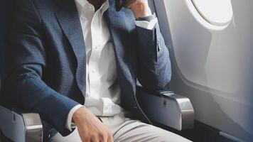 réussi asiatique homme d'affaire dans costume et des lunettes est assis dans privé jet et les usages téléphone intelligent, coréen entrepreneur mouches dans un avion et les types sur le téléphone en ligne video