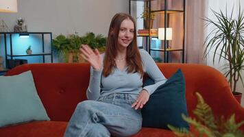 Young woman smiling friendly at camera, waving hands gesturing hello, hi, greeting at home on couch video