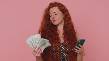 feliz ruiva mulher olhando Smartphone exibição Atenciosamente alegria ganhar, recebendo dinheiro dólar dinheiro video