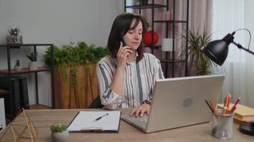 jovem mulher trabalhador autonomo respondendo para cliente em Móvel telefone chamar, usando computador portátil computador, casa escritório video
