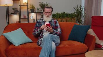 Senior old grandfather sitting on sofa, using smartphone share messages on social media application video