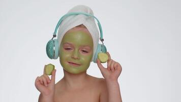 Smiling child girl moisturizing green cucumber mask on face listening to music on headphones dancing video