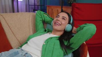 Happy young woman in wireless headphones relaxing lying on sofa at home listening favorite music video