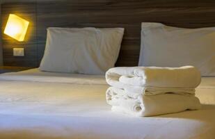 Stack of white towels whick folded neatly on top of bedspread. White bed is neatly made with the covers pulled up tight in hotel room photo
