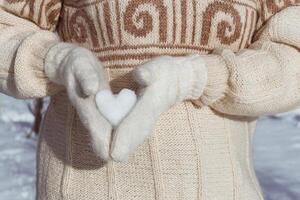 A girl in a beige sweater holds a heart made of snow in her hands, dressed in white mittens, from the side. A gift for Valentine's Day. Postcard with copy space photo