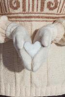 un niña en un beige suéter sostiene en su manos, vestido en blanco mitones, un corazón hecho de nieve de cerca. tarjeta postal para San Valentín día foto