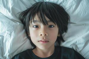 retrato de un 10 año antiguo japonés niño acostado en el cama, mirando a el cámara desde directamente arriba, con asustado cara y melancólico estado animico foto