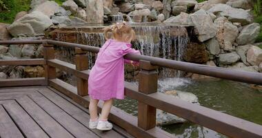 Little child girl in stylish pink dress in summer park and looking at waterfall video