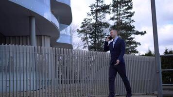 Businessman confidently strides forward, phone pressed to ear. As the Businessman negotiates, office building looms behind. Each step of the Businessman conveys authority and focus video