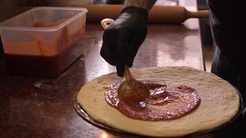 Chef wearing black gloves spreads red tomato sauce with a metal spoon on a pizza base on the table. Concept of cooking delicious pizza. Step by step. Step 2. Middle shot. Slow motion video