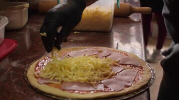 Professional chef wearing black gloves pours grated gouda cheese onto a pizza base with tomato paste. Concept of cooking delicious pizza. Step by step. Step 5. Middle shot. Slow motion video